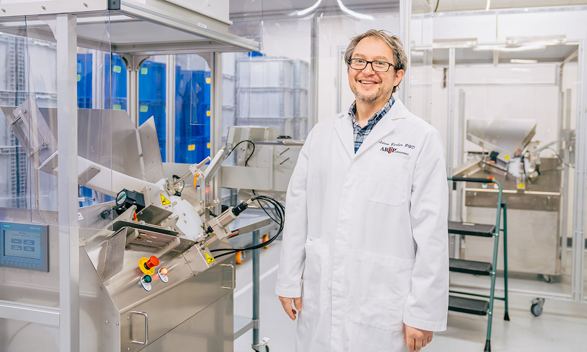 Adam Baker stands in a laboratory. He smiles at the camera.